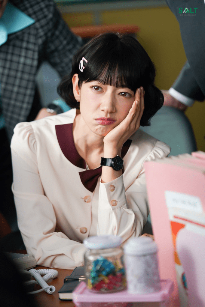 A young woman with short black hair and a pink clip sitting at a desk, resting her chin on her hand with a bored expression. She is wearing a cream-colored blouse with a maroon bow and has a black watch on her wrist. A jar of colorful items and documents are visible on the desk.