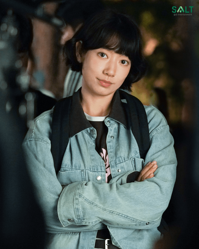 A young woman with short black hair wearing a denim jacket and a black shirt, standing with her arms crossed and looking directly at the camera.