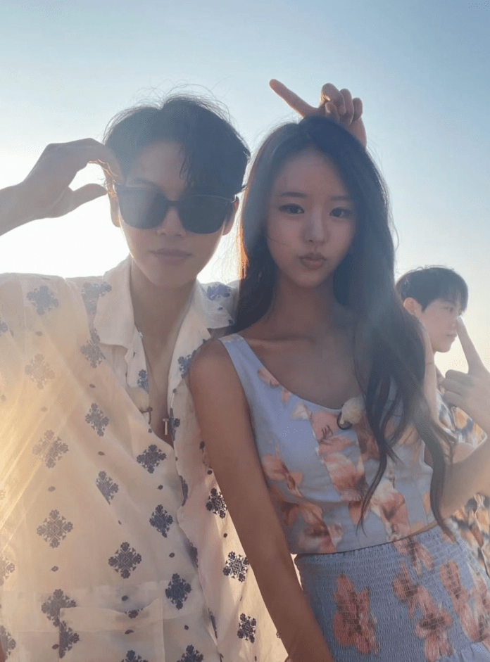 A young man wearing sunglasses and a patterned shirt poses with a young woman in a floral top, both smiling brightly against a sunset background. Another person is partially visible behind them.