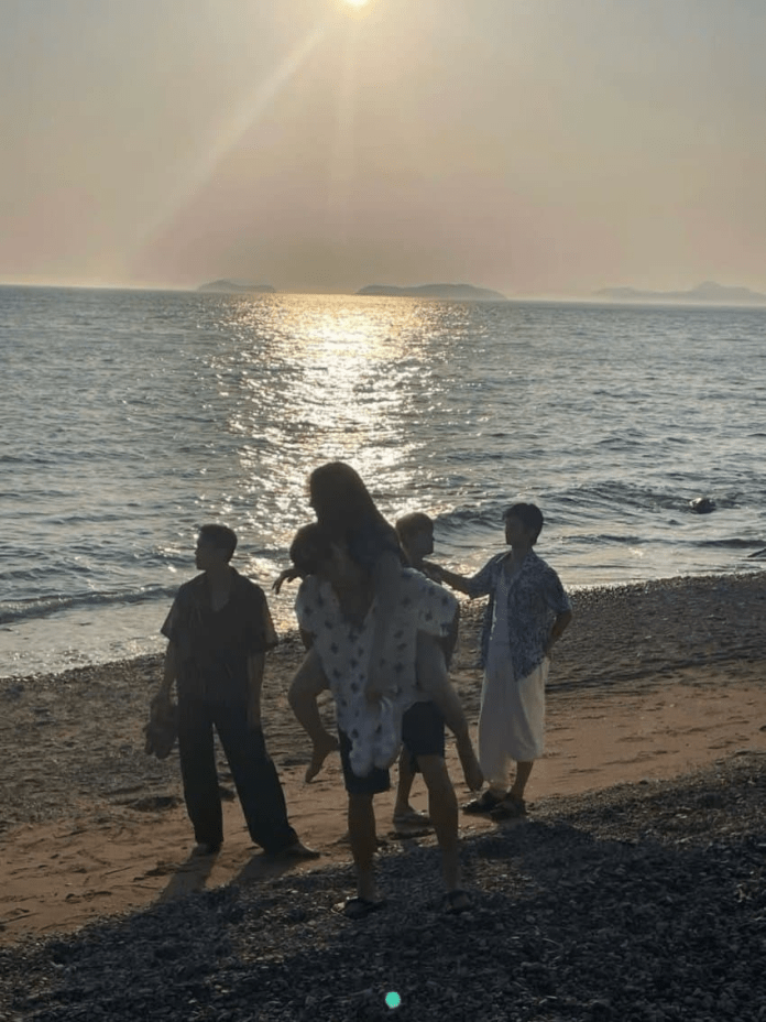 A group of five people standing on a beach during sunset, with ocean waves in the background and silhouettes of distant islands visible.