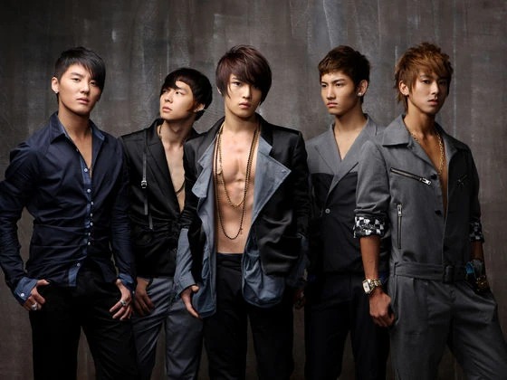 A group of five young men posing together in stylish outfits, set against a neutral background. They have varied hairstyles and are standing confidently, showcasing a modern fashion aesthetic.
