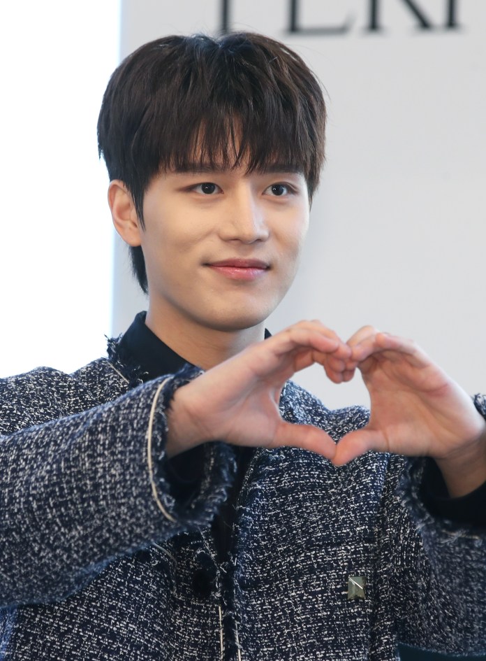 A young man with short dark hair smiling and making a heart shape with his hands.
