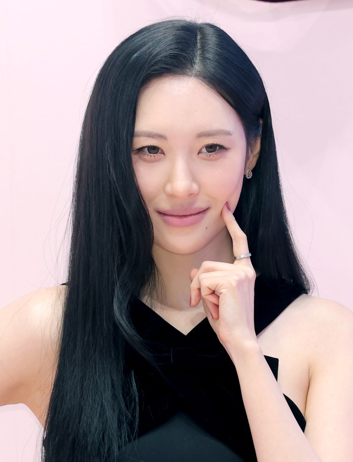 A woman with long black hair smiles while resting her finger on her chin, wearing a black outfit against a soft pink background.