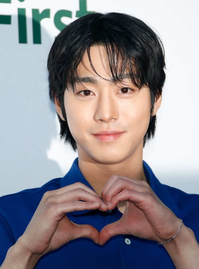A young man with dark hair wearing a blue shirt makes a heart shape with his hands, smiling at the camera.