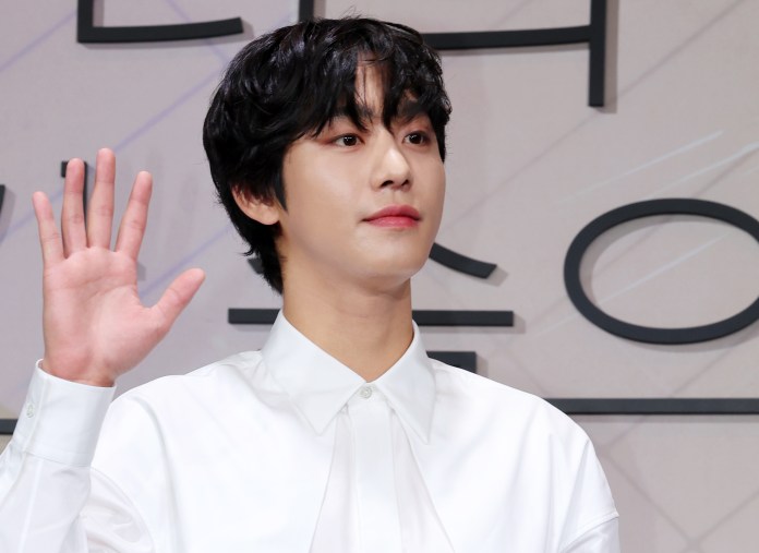 A young man with curly black hair wearing a white shirt is raising his right hand, smiling slightly, against a minimalistic background.