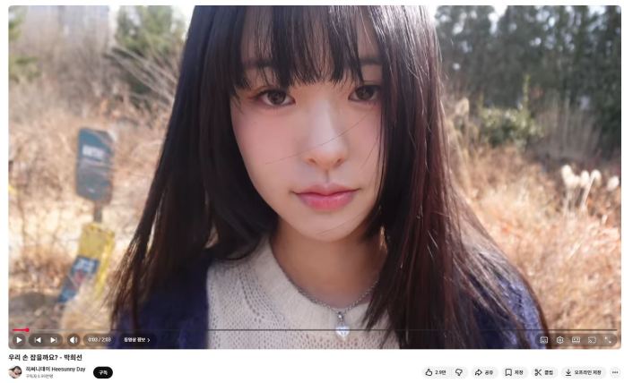 Close-up portrait of a young woman with long dark hair, wearing a light sweater, in a natural outdoor setting with tall grasses in the background.