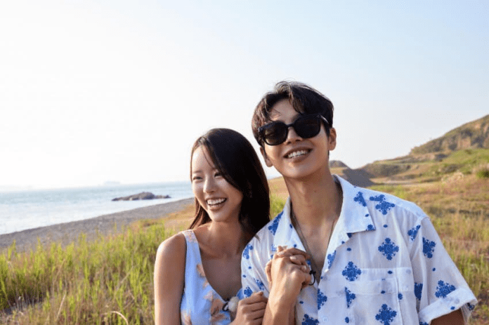 A young couple smiling and holding hands on a beach, with a serene ocean and grassy landscape in the background.