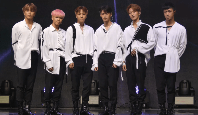 A group of six male performers standing in a row on stage, all wearing black pants and white shirts, with varying styles and accessories.