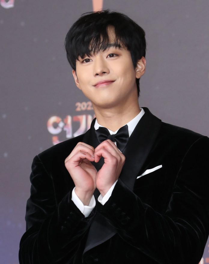 A young man in a black tuxedo smiles and makes a heart shape with his hands at an event.