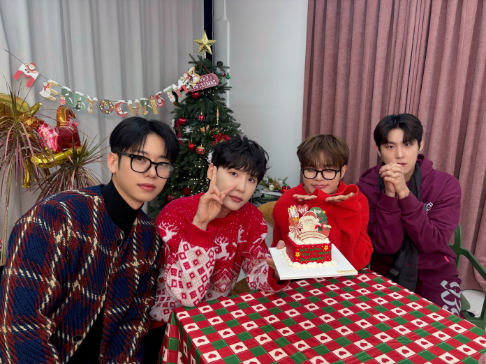 Four friends in festive sweaters pose around a decorated table with a Christmas cake, set against a backdrop of a Christmas tree and holiday decorations.