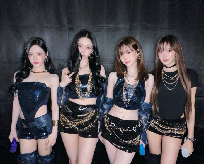 aespa in coordinated sparkly outfits with chains, posing in front of a dark background.