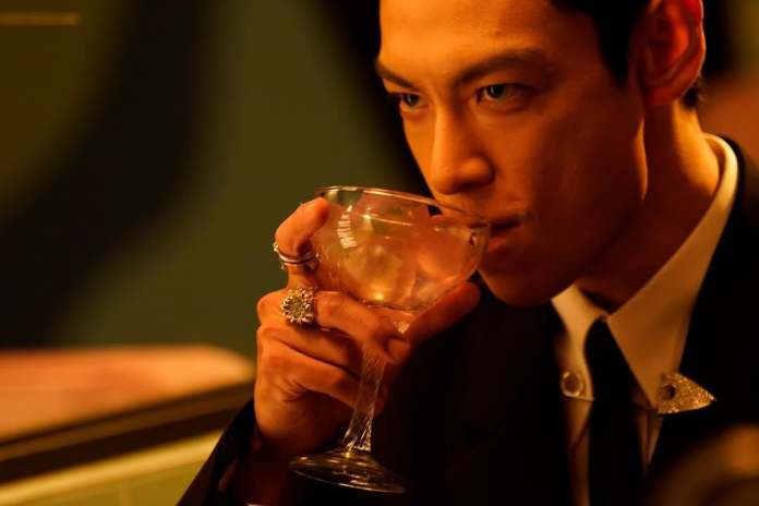 A man in formal attire sipping from a cocktail glass, with a focused expression in a dimly lit setting.