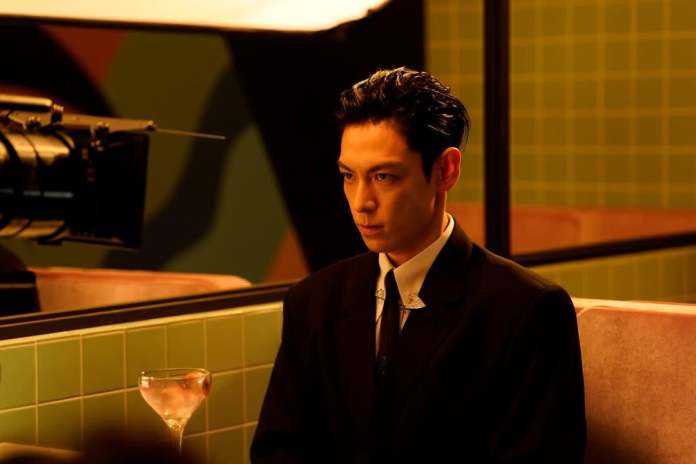 A man in a dark suit sitting at a table with a cocktail, looking intensely in front of him, with a film camera pointed toward him in a stylish, modern setting.