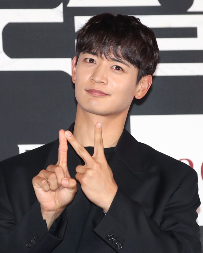 A young man with dark hair and a stylish outfit is smiling and making a peace sign with his hands against a solid backdrop.