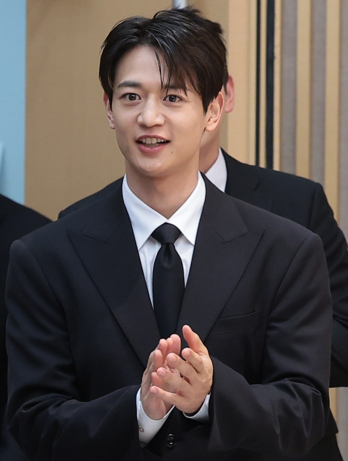 A young man in a black suit and tie claps his hands while smiling, standing in front of a light-colored background with blurred figures in the background.