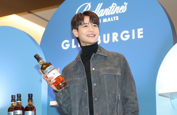 A smiling individual holding a bottle of Ballantine's whiskey, standing in front of a blue background featuring the brand name and Glenburgie.