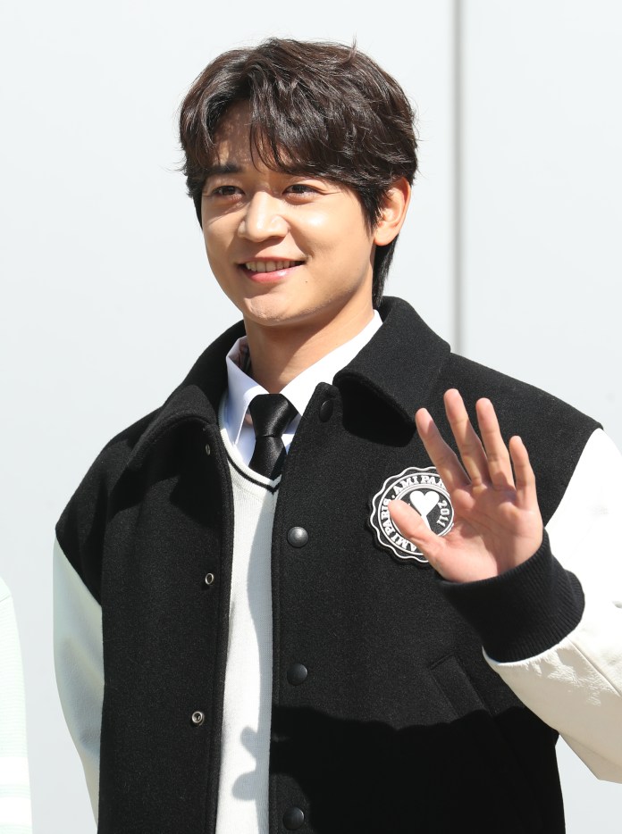 A smiling man in a black and white varsity jacket waves. He has short, styled hair and is wearing a tie over a white sweater.
