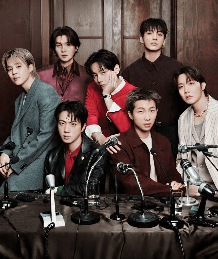 A group of eight men posing together at a table with microphones, dressed in fashionable attire against a wooden backdrop.