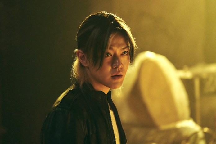 Yuta with long hair, wearing a black jacket, gazes intently in a dimly lit environment.