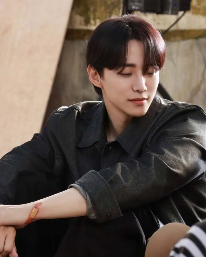 A young person with dark hair wearing a black jacket, sitting with a contemplative expression, and a small bandage on their arm.