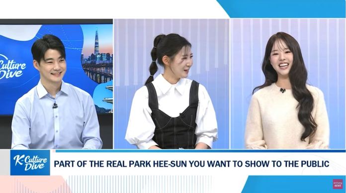 Three hosts smiling and engaging in conversation on a talk show set with a blue and white background, discussing aspects of culture.