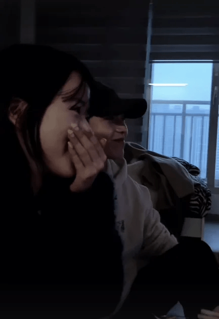 A woman with long dark hair covering her mouth, smiling in surprise while watching something off-screen, sitting next to a man wearing a cap who is also smiling.