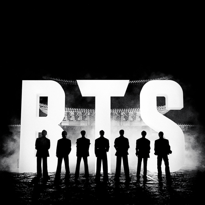 Silhouettes of seven individuals standing in front of a large illuminated sign that reads 'BTS', with a dark background and mist creating a dramatic effect.