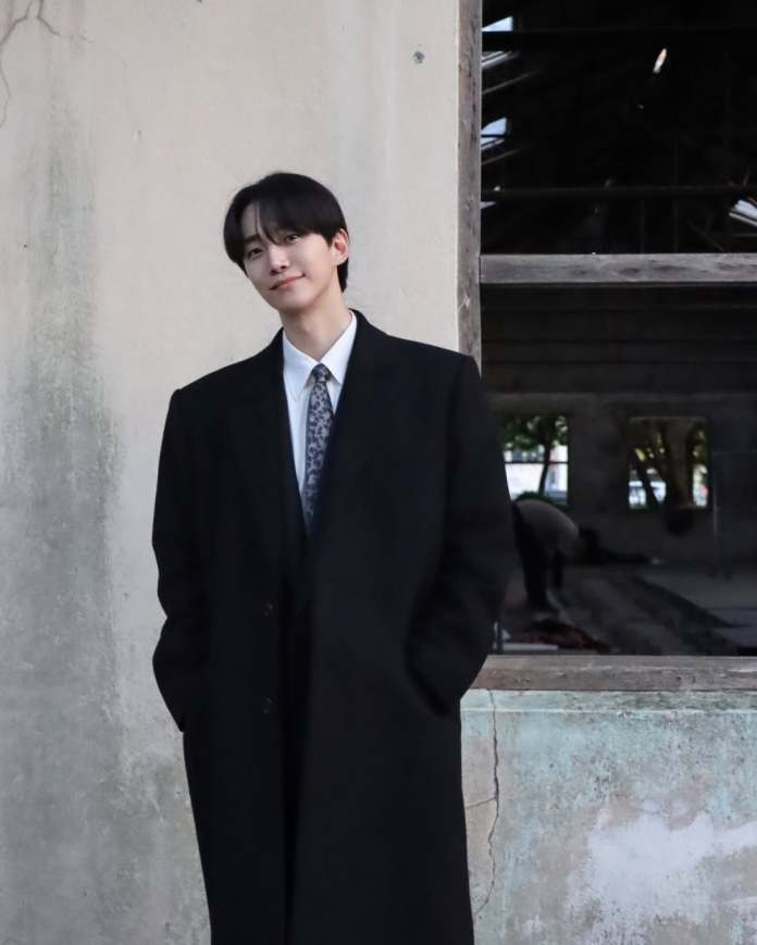 A person wearing a black coat and a white shirt stands with their hands in their pockets, smiling slightly against a weathered wall with a window in the background.