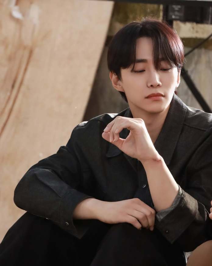 A person sitting quietly with eyes closed, wearing a black jacket, and resting a hand thoughtfully on their knee against a blurred background.