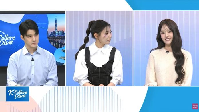 A television show segment featuring three hosts discussing K-culture. The male host on the left is wearing a blue shirt, the female host in the middle is dressed in a black outfit, and the female host on the right is wearing a cream sweater. The background is a soft gradient with the show's logo visible.