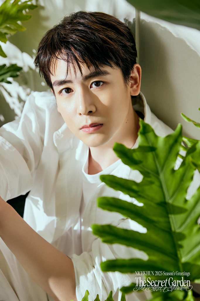 A close-up portrait of a young man with dark hair, wearing a white shirt, sitting among green leafy plants. He has a serious expression and is looking directly at the camera.