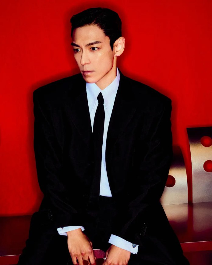 T.O.P wearing a black suit and tie, seated against a vibrant red background, looking thoughtfully to the side.