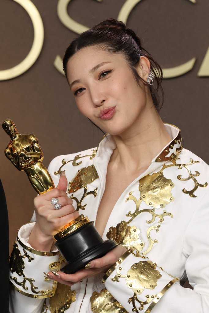 EJAE in an ornate white and gold outfit smiling and holding an Oscar trophy.