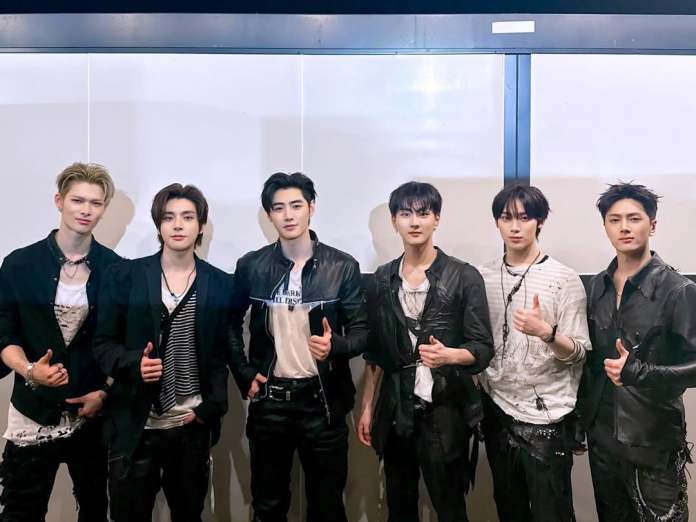 ENHYPEN posing for a photo, all wearing stylish black and white outfits. They are standing in a line, with some giving thumbs up gestures. The background is a plain wall.