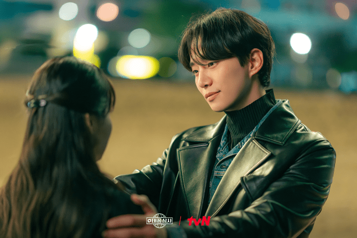 A man wearing a leather jacket and turtleneck gazes at a woman as he holds her hand, set in a softly lit outdoor environment.