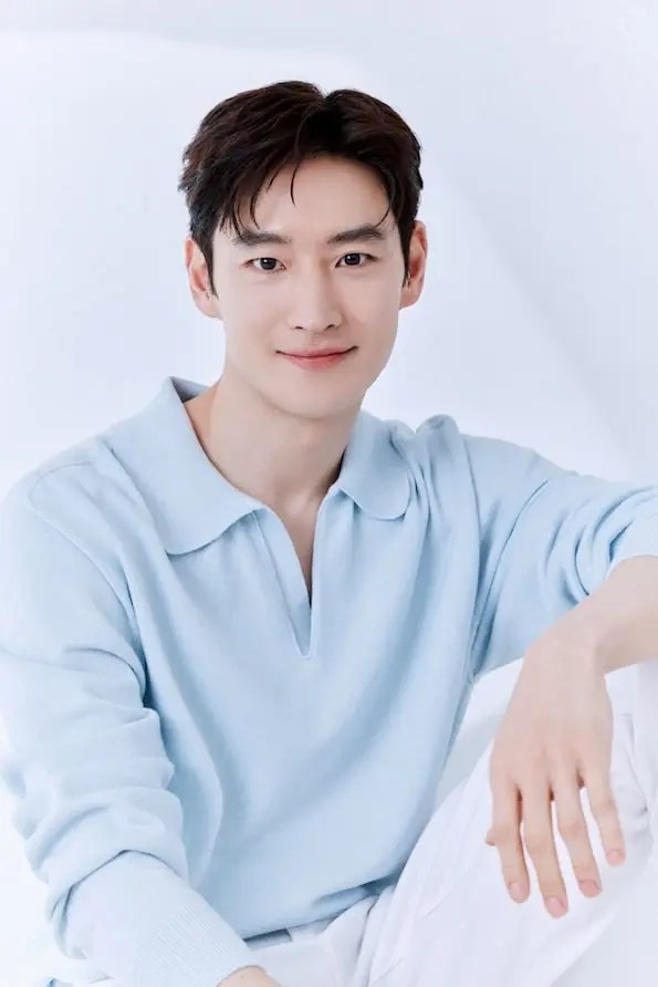 Lee Jehoon with dark hair smiles at the camera while wearing a light blue polo shirt, seated against a soft, white background.