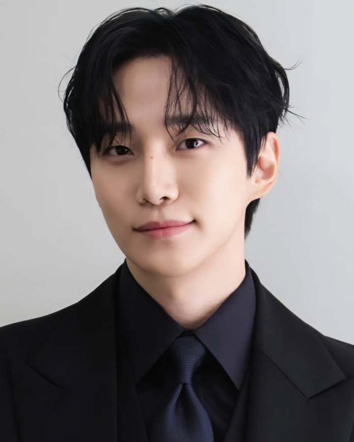 Close-up portrait of a young man wearing a black suit and tie, with dark hair styled softly, set against a light background.