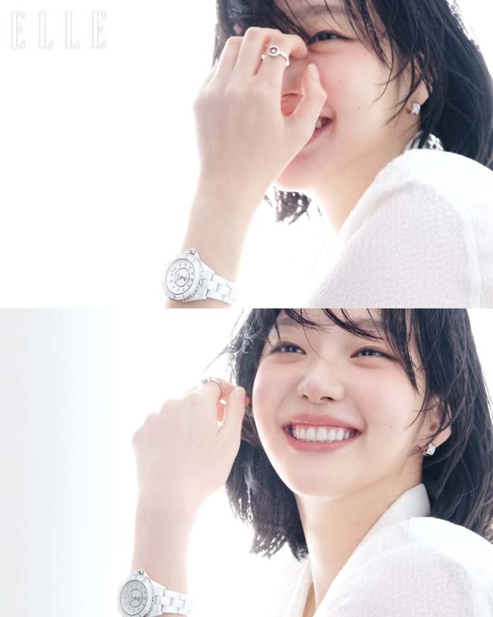Kim Go-eun smiling with short, wet hair, wearing a white top and silver jewelry, gently touches her face while showing a wristwatch against a bright, light background.