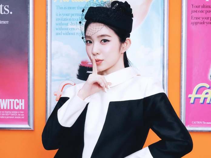 Irene in a stylish black and white outfit poses playfully with a finger to her lips, against a backdrop of colorful posters.
