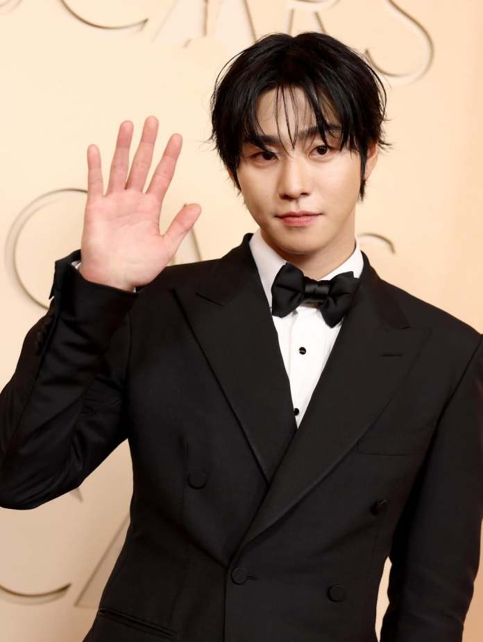 A man in a formal black tuxedo with a bow tie raises his hand in a wave, smiling into the camera against a beige background.
