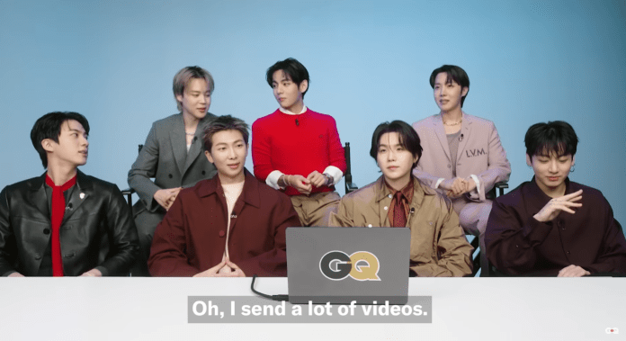BTS seated in a casual interview setting, where one person is speaking about sending videos. The background is light blue, and the group is dressed in stylish attire.