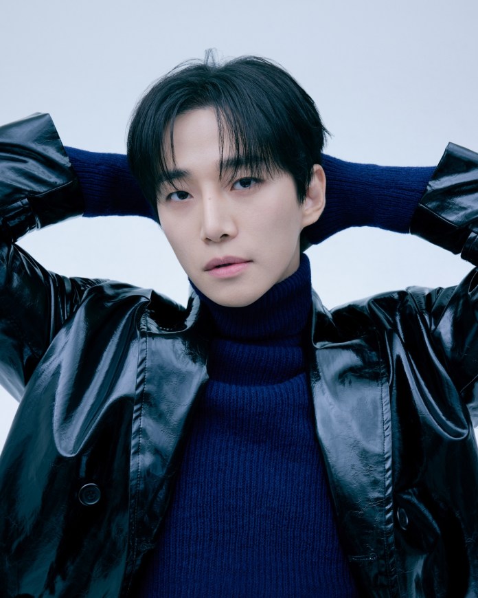 A close-up portrait of a person wearing a glossy black coat and a dark turtleneck sweater, with their hands raised behind their head against a light blue background.