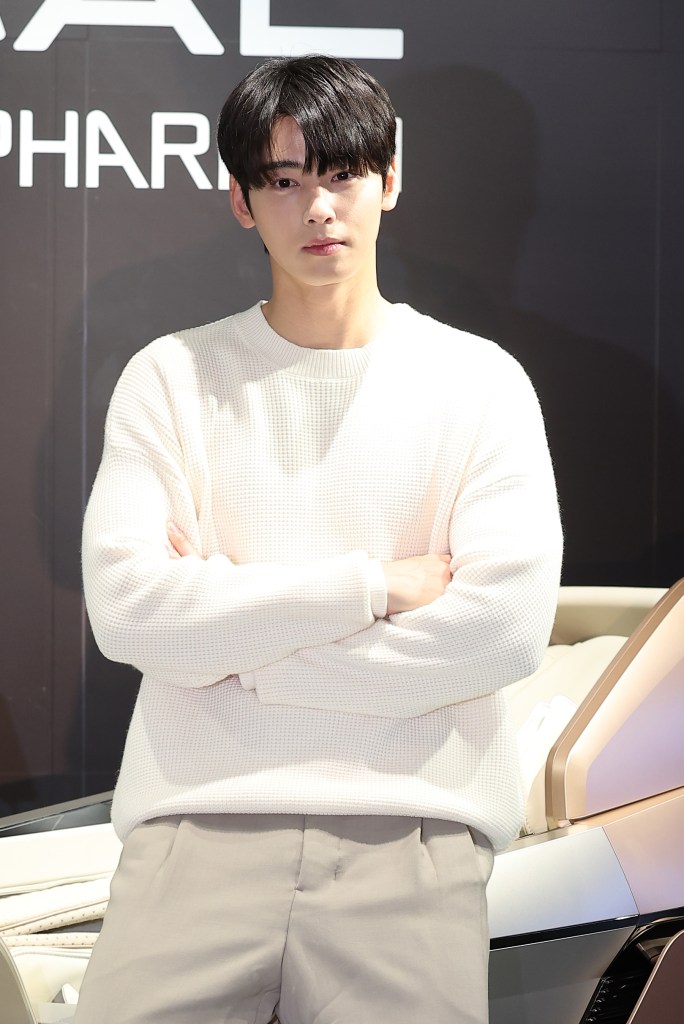 Cha Eun Woo with dark hair wearing a white sweater poses with his arms crossed in front of a modern vehicle, set against a dark backdrop with the word 'PHARMA' visible.