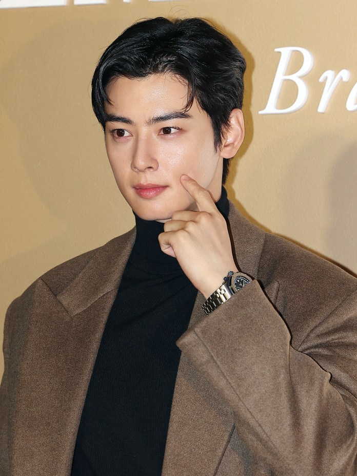 Cha Eun Woo with black hair wearing a brown coat and a black turtleneck, posing and pointing to his face in front of a beige background.