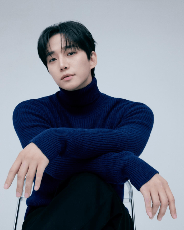 A person sitting on a chair, wearing a navy blue turtleneck sweater, with hands resting casually on their knees and an expressive gaze.