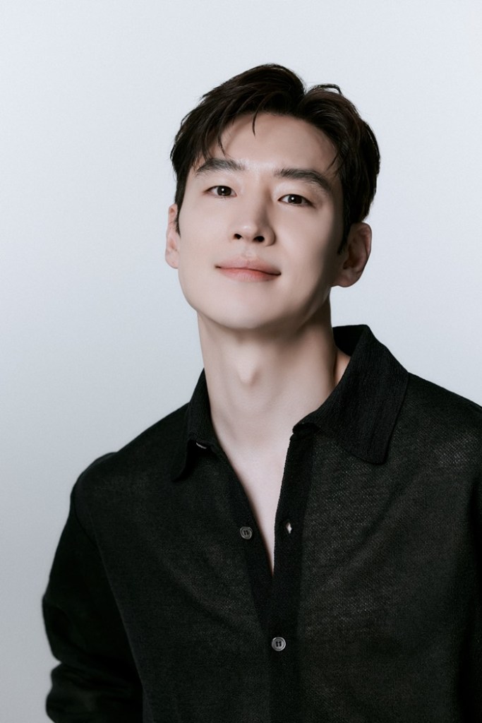Lee Jehoon with short dark hair smiles confidently at the camera, wearing a black button-up shirt against a light background.