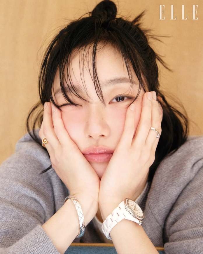 Kim Go-eun is resting their face on their hands, with one eye closed in a playful wink. They are wearing a gray sweater and adorned with a silver bracelet and a white watch. The background is a neutral wooden tone, and the top corner features the word 'ELLE'.