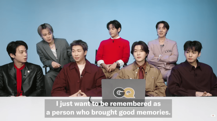 BTS seated at a table during an interview, with one man speaking to the camera. The background is light blue, and text overlay reads, 'I just want to be remembered as a person who brought good memories.'