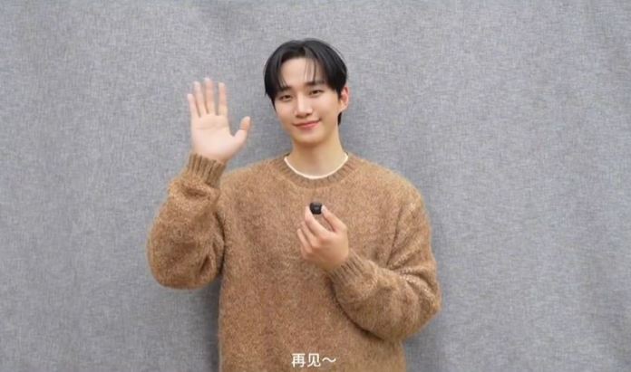 Person in a cozy brown sweater waving hand and smiling against a gray background.