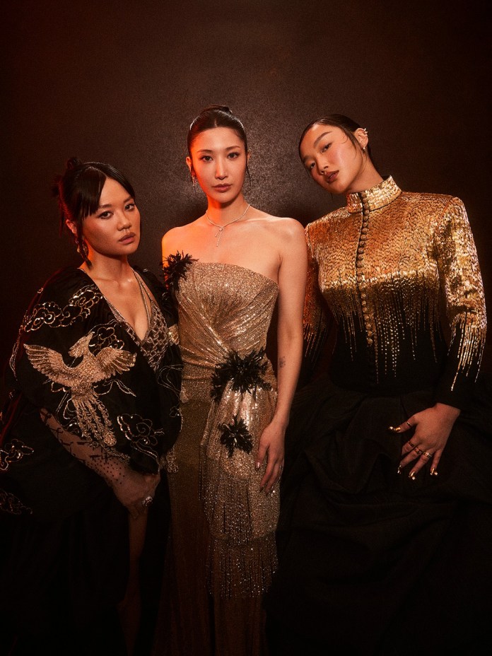 REI AMI, EJAE & AUDREY NUNA pose together in elegant evening wear against a dark background, showcasing various styles and textures in their outfits.